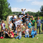 Eine Gruppe von Kindern posiert an einem sonnigen Tag im Freien vor Zielscheiben für das Bogenschießen, einige halten Spielzeugschwerter und zeigen die Daumen nach oben.