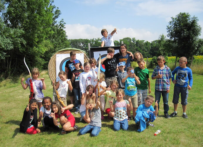 Eine Gruppe von Kindern posiert an einem sonnigen Tag im Freien vor Zielscheiben für das Bogenschießen, einige halten Spielzeugschwerter und zeigen die Daumen nach oben.