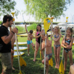 Eine Gruppe von Kindern in Badeanzügen versammelt sich um einen Lehrer mit Paddeln an einem See mit Paddelbrettern und Booten im Hintergrund.