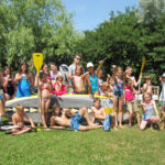 Eine Gruppe von Kindern und ein paar Erwachsene in Badeanzügen posieren im Freien mit Paddeln und einem großen Paddelbrett auf einer Wiese, mit Bäumen im Hintergrund.