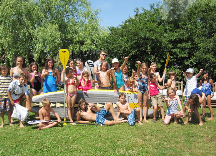 Eine Gruppe von Kindern und ein paar Erwachsene in Badeanzügen posieren im Freien mit Paddeln und einem großen Paddelbrett auf einer Wiese, mit Bäumen im Hintergrund.