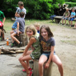 Eine Gruppe von Kindern sitzt im Freien auf Baumstämmen und Bänken in einem Waldgebiet, wobei zwei Mädchen im Vordergrund in die Kamera lächeln.