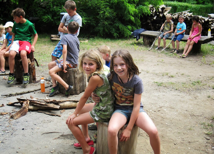 Eine Gruppe von Kindern sitzt im Freien auf Baumstämmen und Bänken in einem Waldgebiet, wobei zwei Mädchen im Vordergrund in die Kamera lächeln.