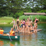Eine Gruppe von Kindern in Badeanzügen steht auf einer schwimmenden Plattform, während andere mit Schwimmwesten in einem Kanu auf einem von Grün umgebenen See paddeln.