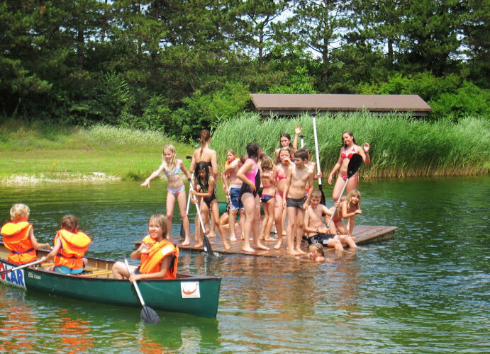 Eine Gruppe von Kindern in Badeanzügen steht auf einer schwimmenden Plattform, während andere mit Schwimmwesten in einem Kanu auf einem von Grün umgebenen See paddeln.