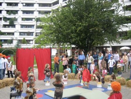 Kinder in Kostümen spielen auf einem Spielplatz im Freien vor einem Publikum, das zuschaut; im Hintergrund sind Wohnhäuser und Bäume zu sehen.