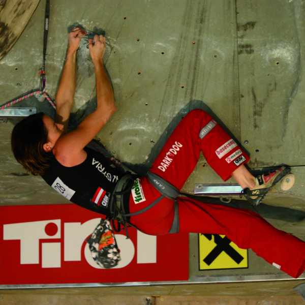 Ein Kletterer in roter Hose und schwarzem Hemd hält sich während eines Wettkampfs an einer Indoor-Kletterwand fest, gesichert mit einem Klettergurt und einem Seil.