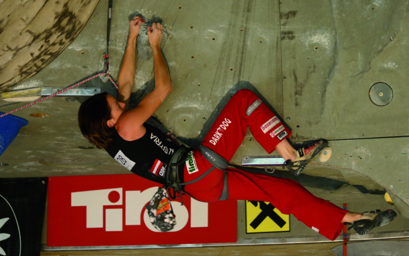 Ein Kletterer in roter Hose und schwarzem Hemd hält sich während eines Wettkampfs an einer Indoor-Kletterwand fest, gesichert mit einem Klettergurt und einem Seil.