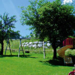 Ein Spielplatz mit Schaukeln, einer gelben Spiralrutsche und einem Spielgerät befindet sich auf einer Wiese mit Bäumen und einem Feld im Hintergrund unter einem klaren blauen Himmel.