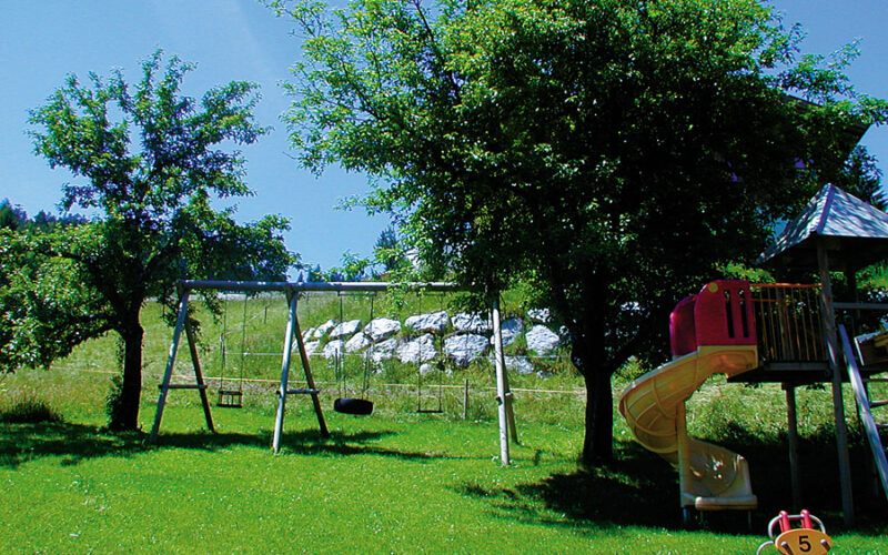 Ein Spielplatz mit Schaukeln, einer gelben Spiralrutsche und einem Spielgerät befindet sich auf einer Wiese mit Bäumen und einem Feld im Hintergrund unter einem klaren blauen Himmel.