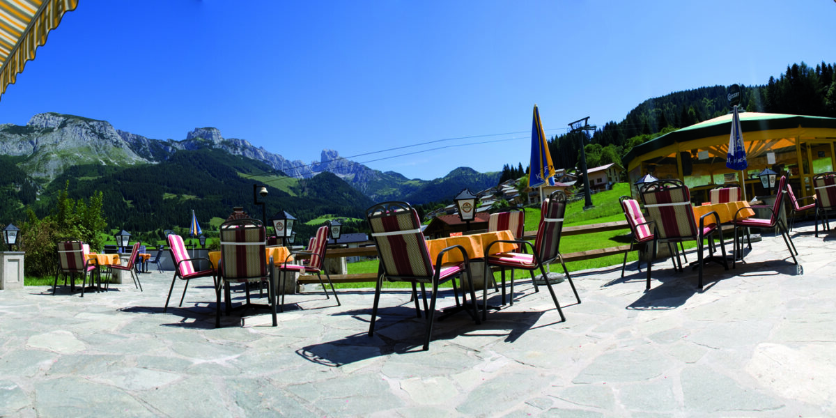 Außencafé mit gestreiften Stühlen und Tischen auf einer Steinterrasse mit Blick auf grüne Hügel und Berge unter einem klaren blauen Himmel.