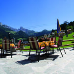 Außencafé mit gestreiften Stühlen und Tischen auf einer Steinterrasse mit Blick auf grüne Hügel und Berge unter einem klaren blauen Himmel.