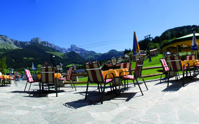 Außencafé mit gestreiften Stühlen und Tischen auf einer Steinterrasse mit Blick auf grüne Hügel und Berge unter einem klaren blauen Himmel.