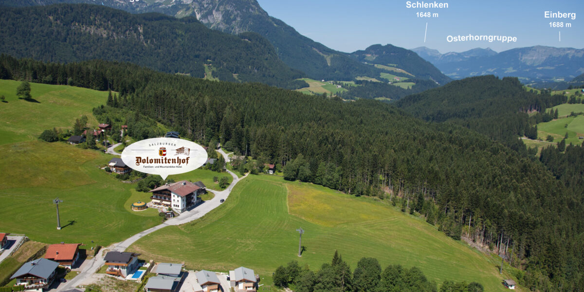 Luftaufnahme eines ländlichen Dorfes, umgeben von grünen Feldern und Wäldern, mit markanten Berggipfeln im Hintergrund unter einem klaren Himmel.