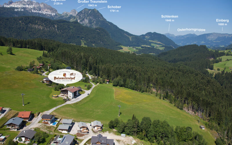 Luftaufnahme eines ländlichen Dorfes, umgeben von grünen Feldern und Wäldern, mit markanten Berggipfeln im Hintergrund unter einem klaren Himmel.