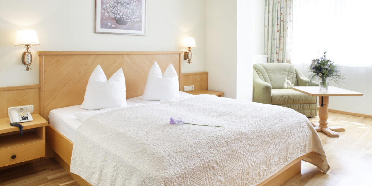 Ein ordentlich gemachtes Hotelbett mit weißer Bettwäsche und Kissen, ein kleiner Tisch mit Stühlen am Fenster und eine Blume auf der Bettdecke in einem hellen, aufgeräumten Zimmer.