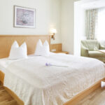 Ein ordentlich gemachtes Hotelbett mit weißer Bettwäsche und Kissen, ein kleiner Tisch mit Stühlen am Fenster und eine Blume auf der Bettdecke in einem hellen, aufgeräumten Zimmer.