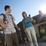 Zwei Personen mit Trekkingstöcken stehen draußen im Sonnenlicht, lächeln sich an, tragen legere Wanderkleidung und Rucksäcke vor einem begrünten Gebäude.