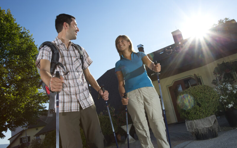 Zwei Personen mit Trekkingstöcken stehen draußen im Sonnenlicht, lächeln sich an, tragen legere Wanderkleidung und Rucksäcke vor einem begrünten Gebäude.