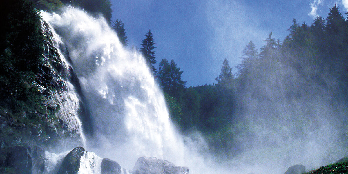Ein Wasserfall stürzt über felsige Klippen, die von immergrünen Bäumen umgeben sind, unter blauem Himmel in die Tiefe, wobei Nebel aus dem fallenden Wasser aufsteigt.