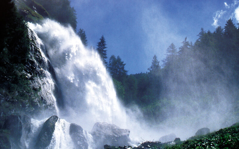 Ein Wasserfall stürzt über felsige Klippen, die von immergrünen Bäumen umgeben sind, unter blauem Himmel in die Tiefe, wobei Nebel aus dem fallenden Wasser aufsteigt.