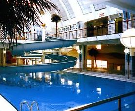 Hallenbad mit spiralförmiger Wasserrutsche, umgeben von Geländern und künstlichen Palmen unter einer hohen, weißen Gewölbedecke.