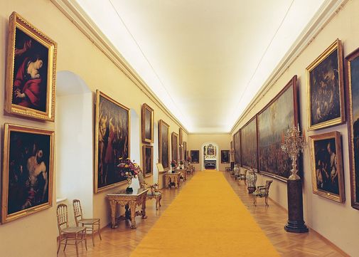 Eine lange, verzierte Galerie mit einem gelben Teppich, gesäumt von gerahmten Bildern an beiden Wänden, Tischen mit Vasen und eleganten Stühlen.