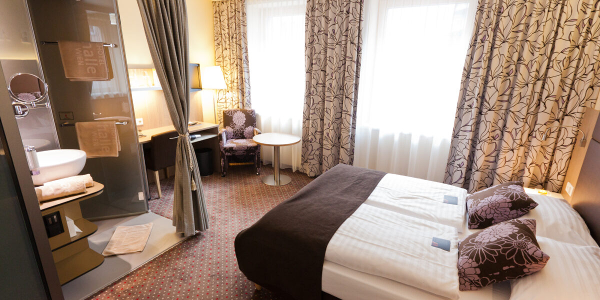 Hotelzimmer mit einem Doppelbett, gemusterten Vorhängen, einem Schreibtisch, einem Stuhl, einem kleinen Tisch und einem offenen Badezimmer mit Glaswänden, Waschbecken und Handtüchern. Natürliches Licht durch zwei große Fenster.