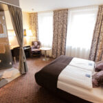 Hotelzimmer mit einem Doppelbett, gemusterten Vorhängen, einem Schreibtisch, einem Stuhl, einem kleinen Tisch und einem offenen Badezimmer mit Glaswänden, Waschbecken und Handtüchern. Natürliches Licht durch zwei große Fenster.