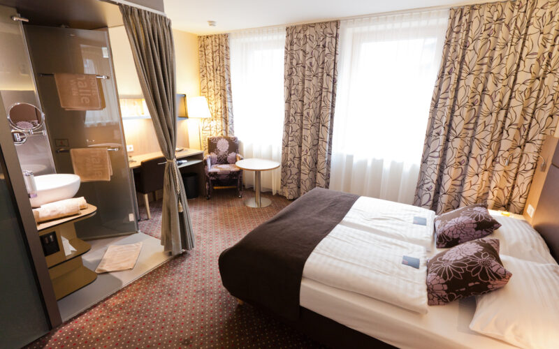 Hotelzimmer mit einem Doppelbett, gemusterten Vorhängen, einem Schreibtisch, einem Stuhl, einem kleinen Tisch und einem offenen Badezimmer mit Glaswänden, Waschbecken und Handtüchern. Natürliches Licht durch zwei große Fenster.