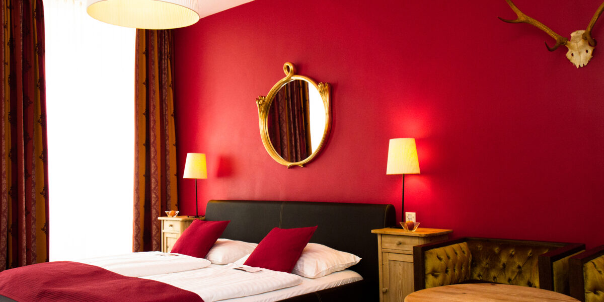Ein Hotelzimmer mit einem Doppelbett, roten Wänden, zwei Nachttischen mit Lampen, einem runden Spiegel, einem Geweih an der Wand und einem runden Holztisch mit einem gepolsterten Sofa.
