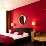 Ein Hotelzimmer mit einem Doppelbett, roten Wänden, zwei Nachttischen mit Lampen, einem runden Spiegel, einem Geweih an der Wand und einem runden Holztisch mit einem gepolsterten Sofa.