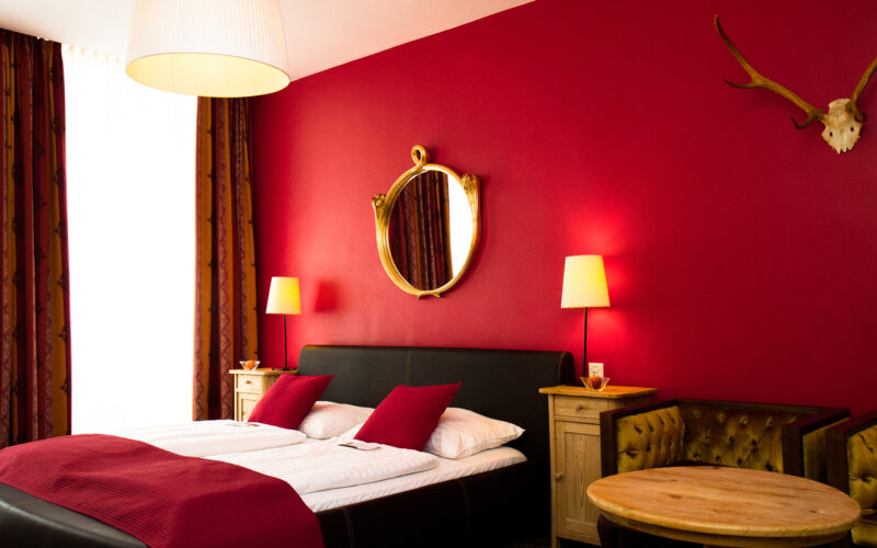 Ein Hotelzimmer mit einem Doppelbett, roten Wänden, zwei Nachttischen mit Lampen, einem runden Spiegel, einem Geweih an der Wand und einem runden Holztisch mit einem gepolsterten Sofa.