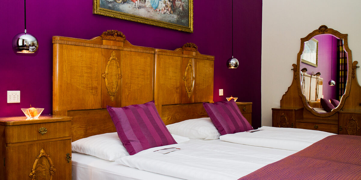Ein Schlafzimmer mit zwei zusammengestellten Einzelbetten, Kopfteilen und Möbeln aus Holz, einer lilafarbenen Akzentwand, passendem Bettzeug und einem dekorativen Gemälde über den Betten.