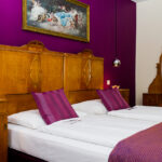 Ein Schlafzimmer mit zwei zusammengestellten Einzelbetten, Kopfteilen und Möbeln aus Holz, einer lilafarbenen Akzentwand, passendem Bettzeug und einem dekorativen Gemälde über den Betten.