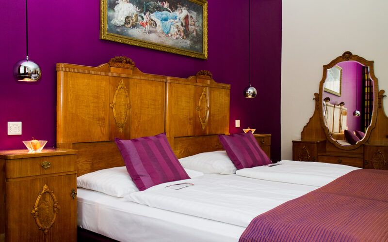 Ein Schlafzimmer mit zwei zusammengestellten Einzelbetten, Kopfteilen und Möbeln aus Holz, einer lilafarbenen Akzentwand, passendem Bettzeug und einem dekorativen Gemälde über den Betten.