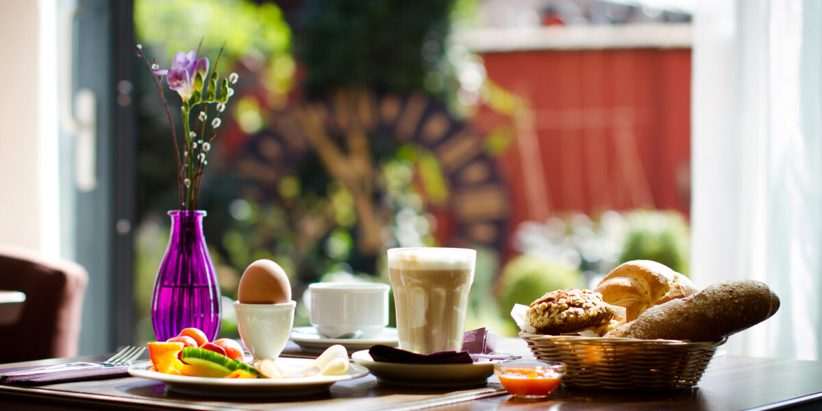 Ein gedeckter Frühstückstisch mit Kaffee, einem gekochten Ei, Brot, Gebäck, Obst und einer violetten Vase mit einer einzelnen Blume, neben einem sonnenbeschienenen Fenster mit Blick auf den Garten.