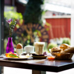 Ein gedeckter Frühstückstisch mit Kaffee, einem gekochten Ei, Brot, Gebäck, Obst und einer violetten Vase mit einer einzelnen Blume, neben einem sonnenbeschienenen Fenster mit Blick auf den Garten.