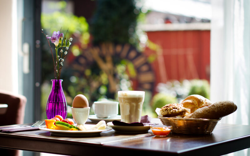 Ein gedeckter Frühstückstisch mit Kaffee, einem gekochten Ei, Brot, Gebäck, Obst und einer violetten Vase mit einer einzelnen Blume, neben einem sonnenbeschienenen Fenster mit Blick auf den Garten.