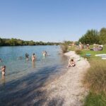 An einem sonnigen Tag baden und entspannen sich die Menschen am grasbewachsenen und kiesigen Ufer eines Sees, wobei Handtücher und Schlauchboote sichtbar sind.