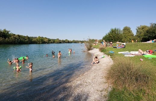 An einem sonnigen Tag baden und entspannen sich die Menschen am grasbewachsenen und kiesigen Ufer eines Sees, wobei Handtücher und Schlauchboote sichtbar sind.