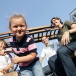 Drei Kinder und zwei Erwachsene sitzen lächelnd auf einer Holzbank im Freien unter strahlend blauem Himmel, im Hintergrund ist ein Gebäude zu sehen.