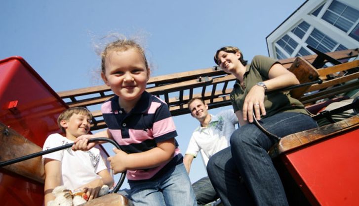 Drei Kinder und zwei Erwachsene sitzen lächelnd auf einer Holzbank im Freien unter strahlend blauem Himmel, im Hintergrund ist ein Gebäude zu sehen.