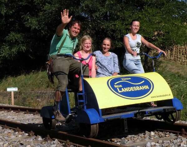 Vier Personen fahren auf einem blau-gelben Schienenfahrrad mit der Aufschrift "Neue Landesbahn" auf Bahngleisen, mit Bäumen und Gras im Hintergrund. Eine Person winkt in die Kamera.