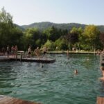 An einem sonnigen Tag schwimmen und spielen mehrere Personen in einem natürlichen Freibad, das von Bäumen, Grasflächen und entfernten Hügeln umgeben ist.