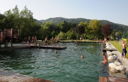 An einem sonnigen Tag schwimmen und spielen mehrere Personen in einem natürlichen Freibad, das von Bäumen, Grasflächen und entfernten Hügeln umgeben ist.