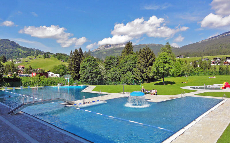 Außenschwimmbad mit Springbrunnen, umgeben von grünen Rasenflächen, Bäumen und Hügeln unter einem teilweise bewölkten Himmel.