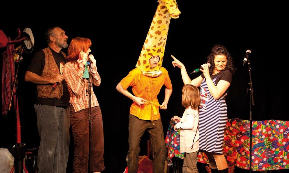Fünf Personen treten auf der Bühne auf; eine trägt ein Giraffenkostüm, andere stehen oder halten Mikrofone, und im Hintergrund steht ein bunter Tisch.
