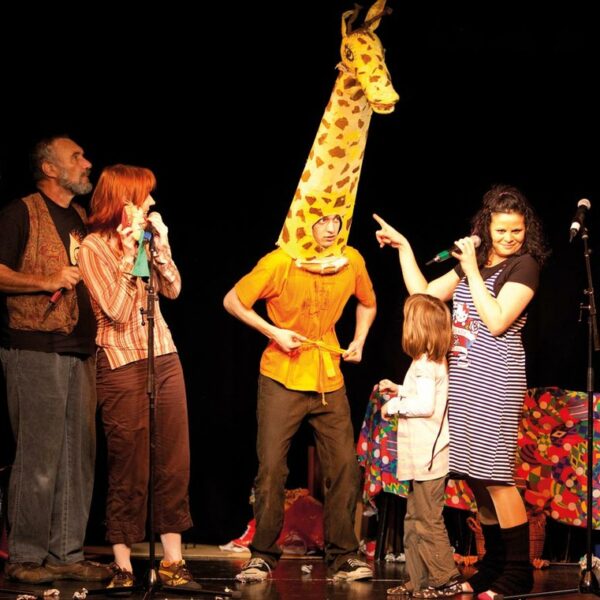 Fünf Personen treten auf der Bühne auf; eine trägt ein Giraffenkostüm, andere stehen oder halten Mikrofone, und im Hintergrund steht ein bunter Tisch.
