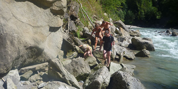 Vier Personen wandern an einem felsigen Flussufer entlang, wobei sie bei Tageslicht über große Steine und Felsbrocken in der Nähe von fließendem Wasser navigieren.